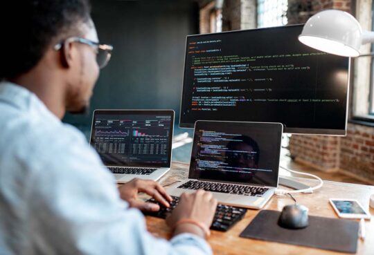 Young african male programmer writing program code sitting at the workplace with three monitors in the office. Image focused on the screen