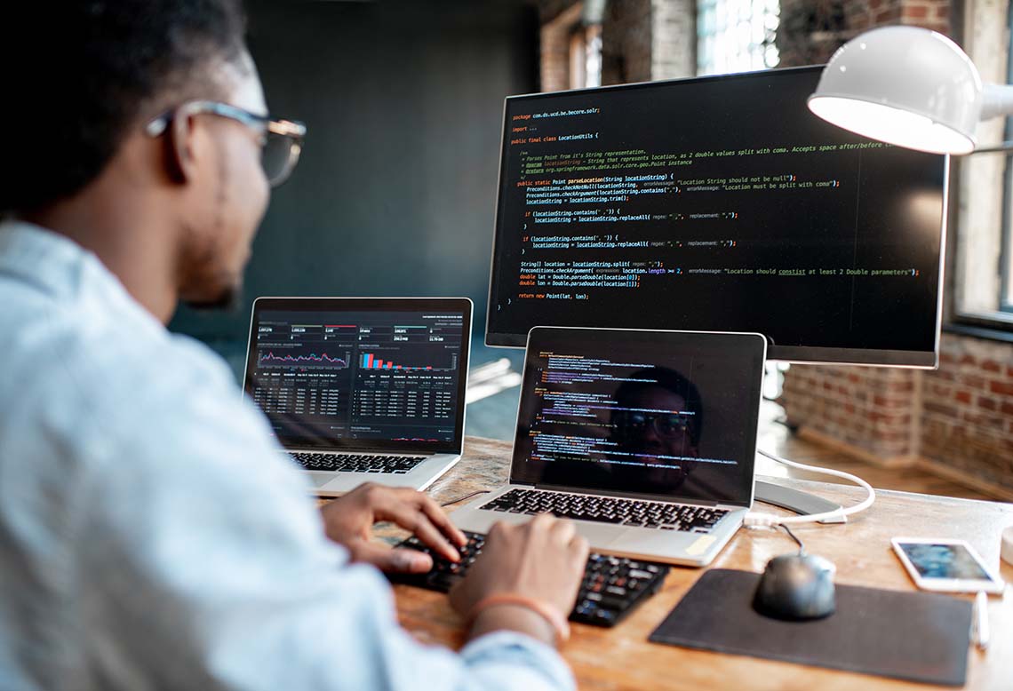 Young african male programmer writing program code sitting at the workplace with three monitors in the office. Image focused on the screen